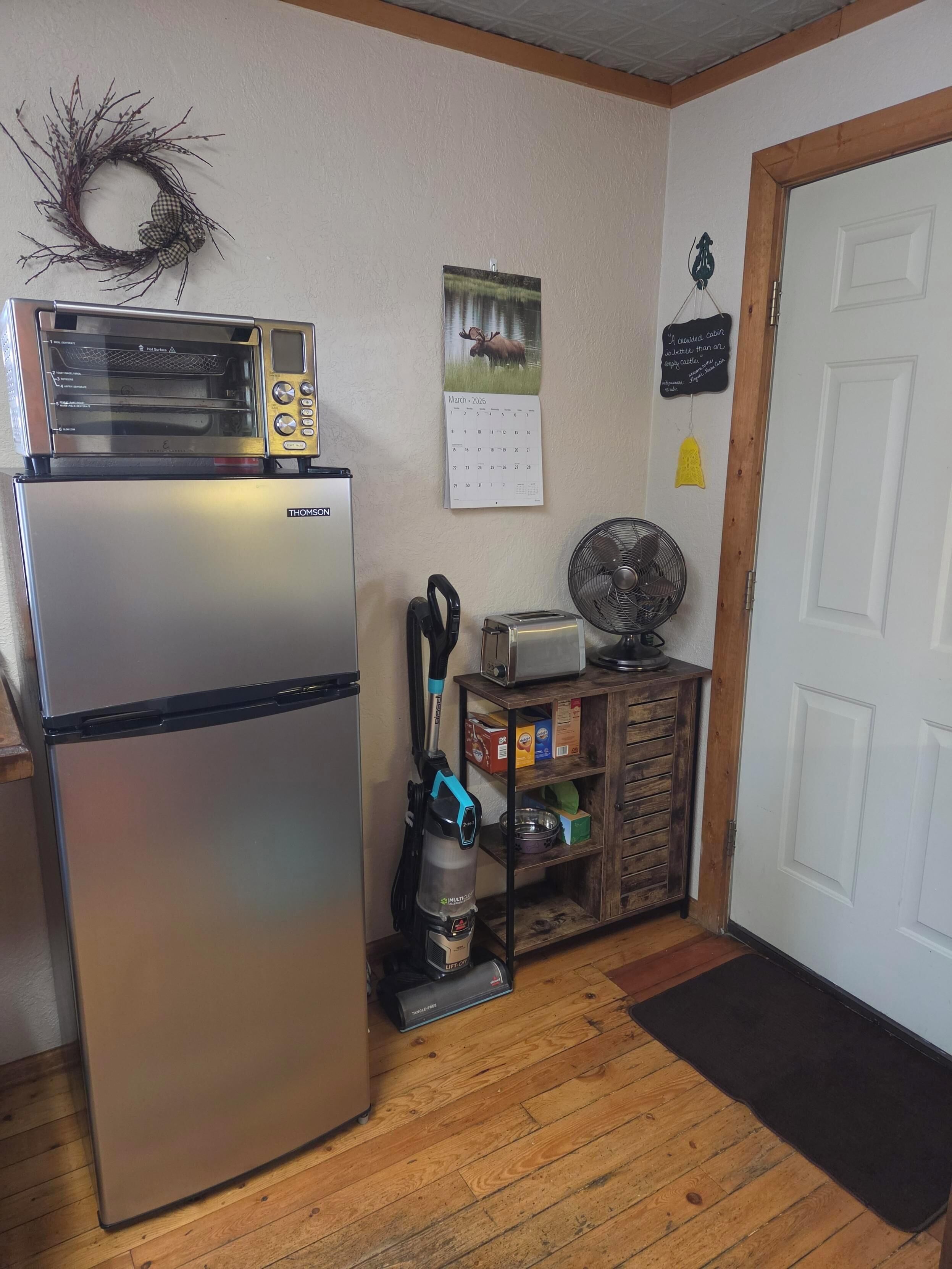 Una cocina con un refrigerador de acero inoxidable, un tostador, una aspiradora, un calendario y un armario de madera con alimentos.