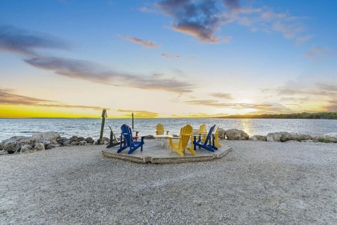Un área circular con asientos, sillas y una mesa sobre un suelo de grava, con vista a un cuerpo de agua y un atardecer al fondo.