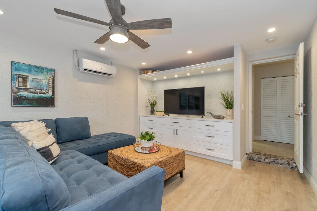 Uma sala de estar moderna com um sofá azul, uma televisão sobre um armário branco e um ventilador de teto.
