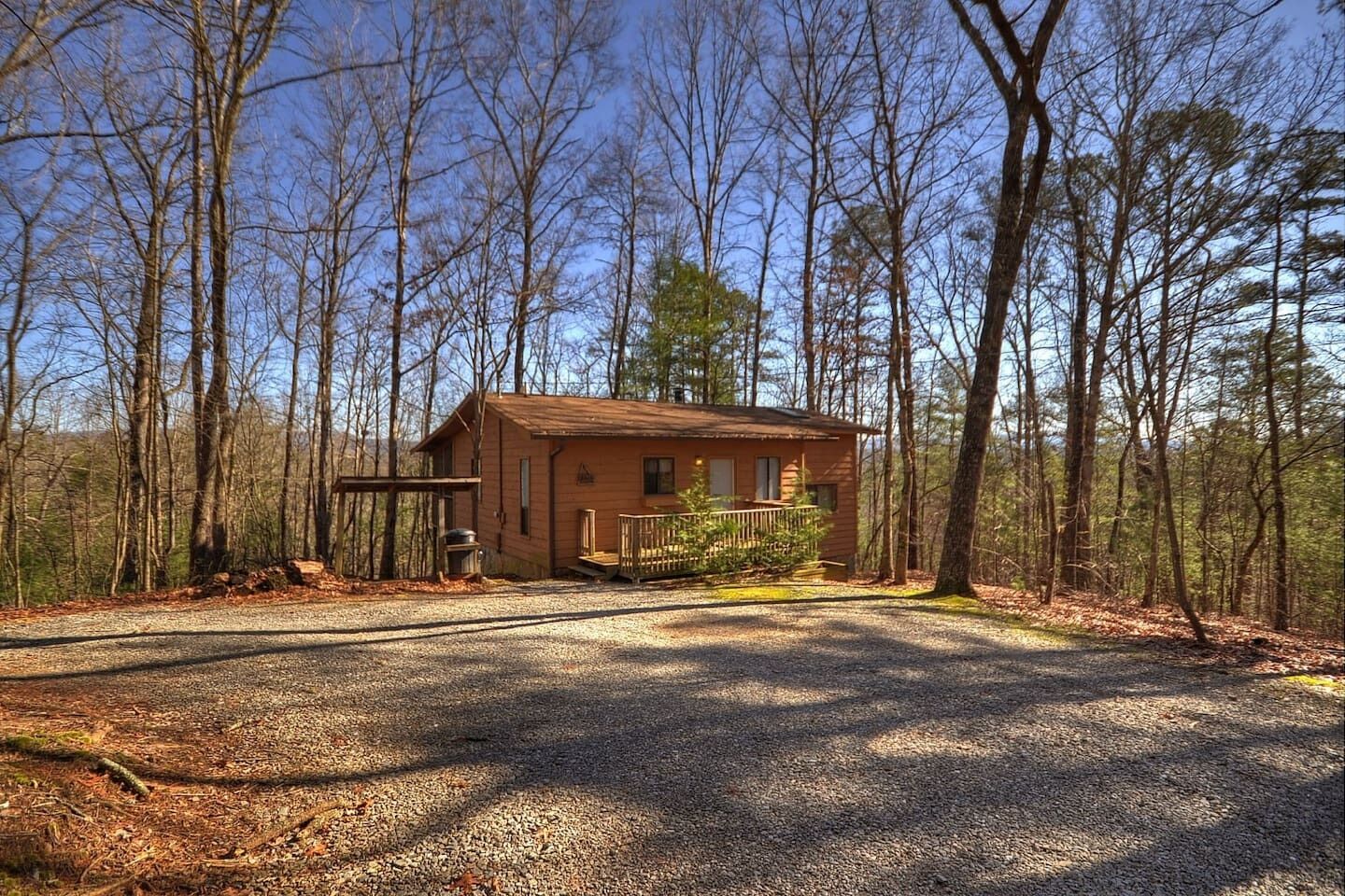 A small cabin with a gravel driveway and a wooden deck, surrounded by trees.