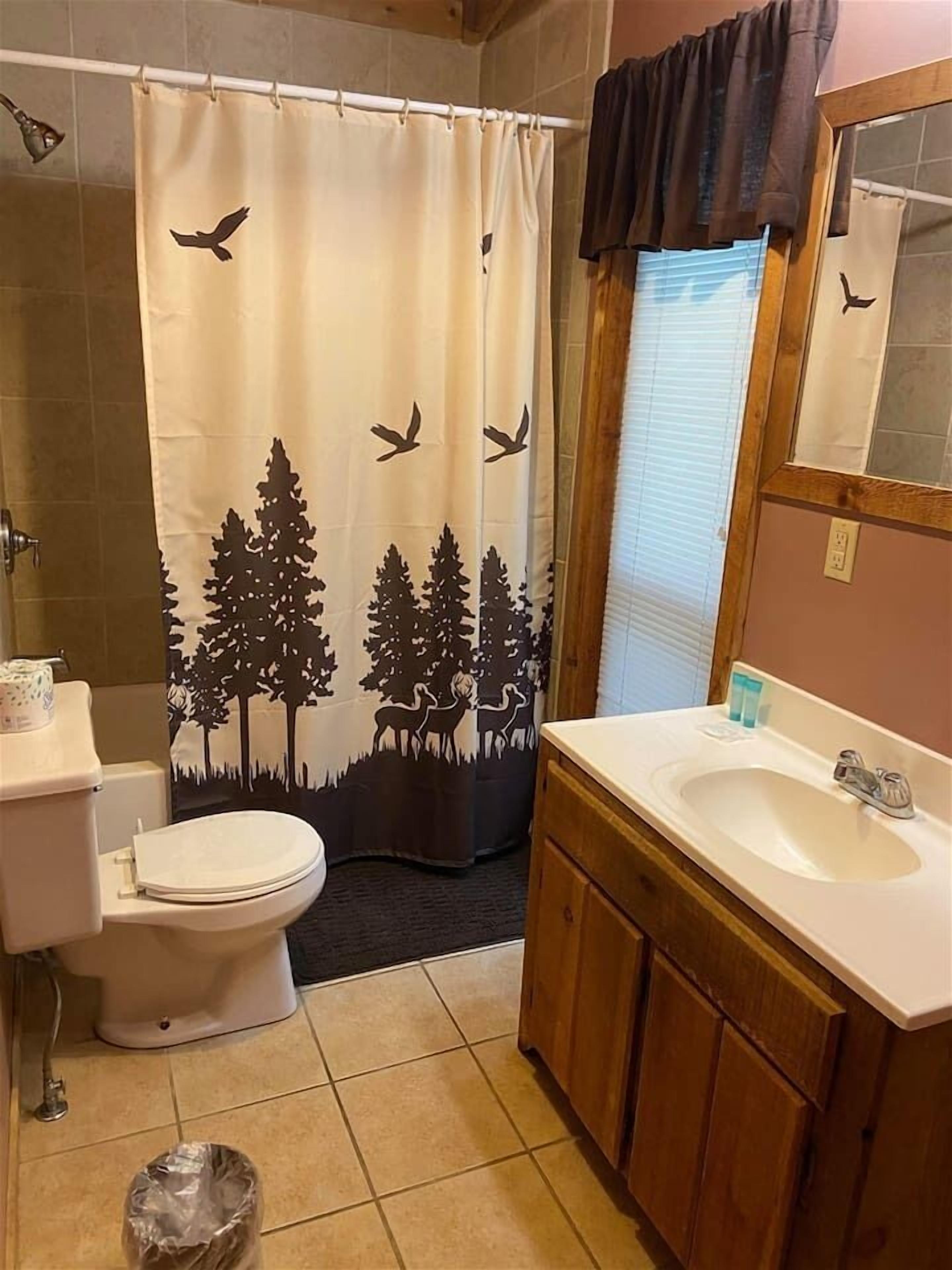 Une salle de bain équipée de toilettes, d’un lavabo et d’un rideau de douche orné d’un motif représentant une forêt et des oiseaux.