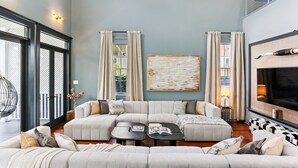 A spacious living room with a large sectional sofa, a coffee table, and a fireplace. There are curtains, a painting, and a large window providing natural light. - Designer Home on Magazine | Pool and Spa (New Orleans)
