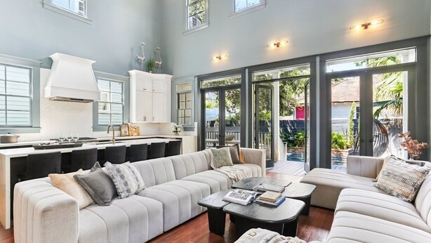A spacious living area with a large sofa and coffee table, a kitchen with a modern design, and large windows providing natural light. - Designer Home on Magazine | Pool and Spa (New Orleans)