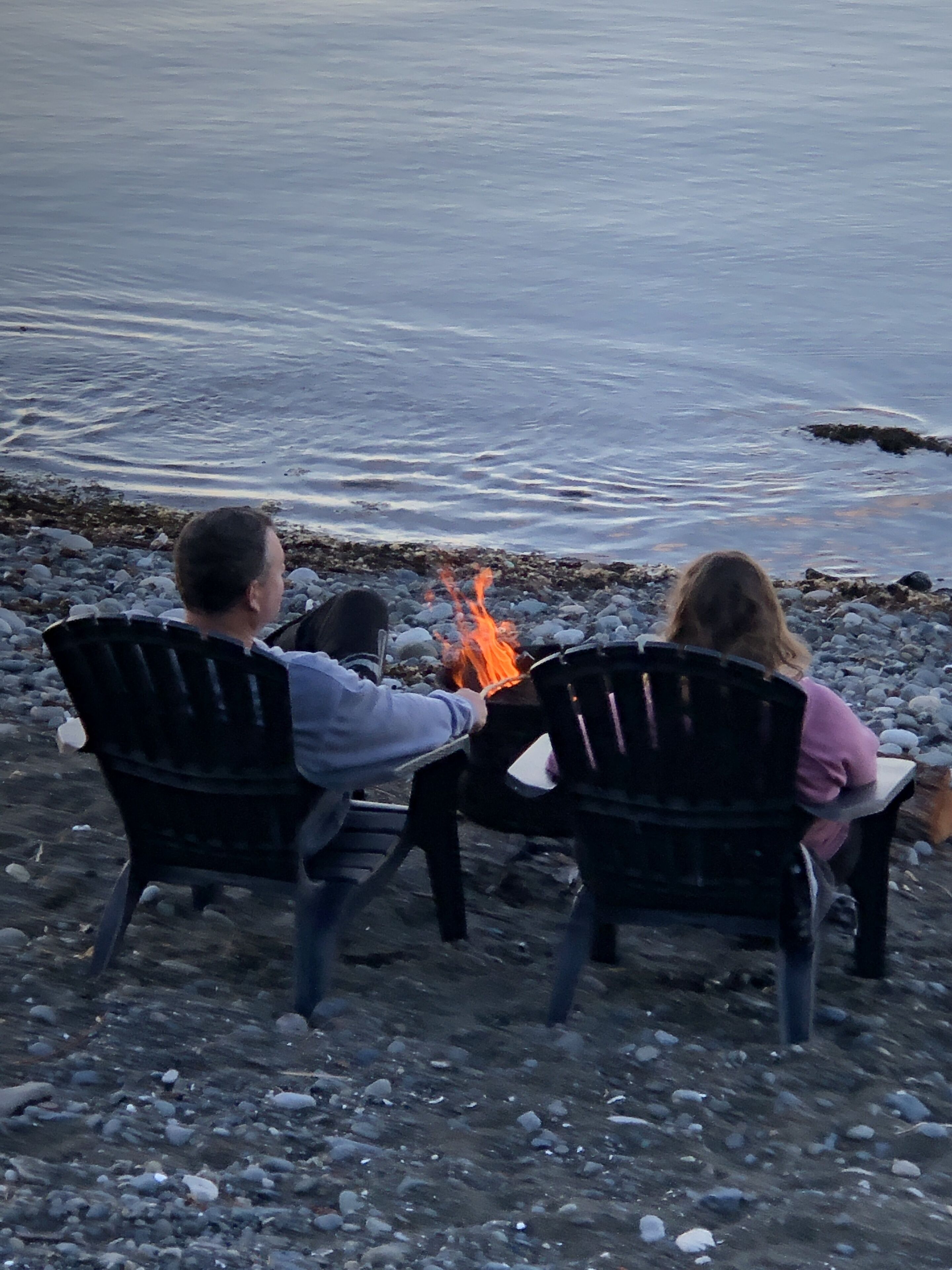 Deux personnes sont assises sur des chaises, face à un feu de camp sur une plage rocheuse, avec une étendue d'eau en arrière-plan.