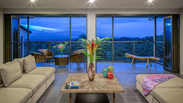 A spacious living area with a large window overlooking a mountainous landscape. There is a sofa, a coffee table with books, and a decorative vase with flowers. - 6BR Luxury Contemporary Villa • Private Pool • Game Room • Quiet Setting (Manuel Antonio)