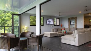 A spacious living room with a large sofa, a coffee table, and wicker chairs. There is a TV mounted on the wall and a large window providing a view of greenery outside.