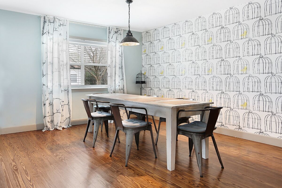 A dining area with a wooden table and four chairs, a window with curtains, and a wall with a birdcage pattern.