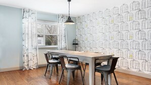 Un comedor con una mesa de madera y cuatro sillas, una ventana con cortinas, y una pared con estampado de jaulas de pájaros.