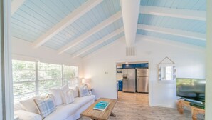 Una sala de estar moderna con un sofá blanco, una mesa de centro de madera y un televisor. Se ve una cocina con electrodomésticos de acero inoxidable al fondo.