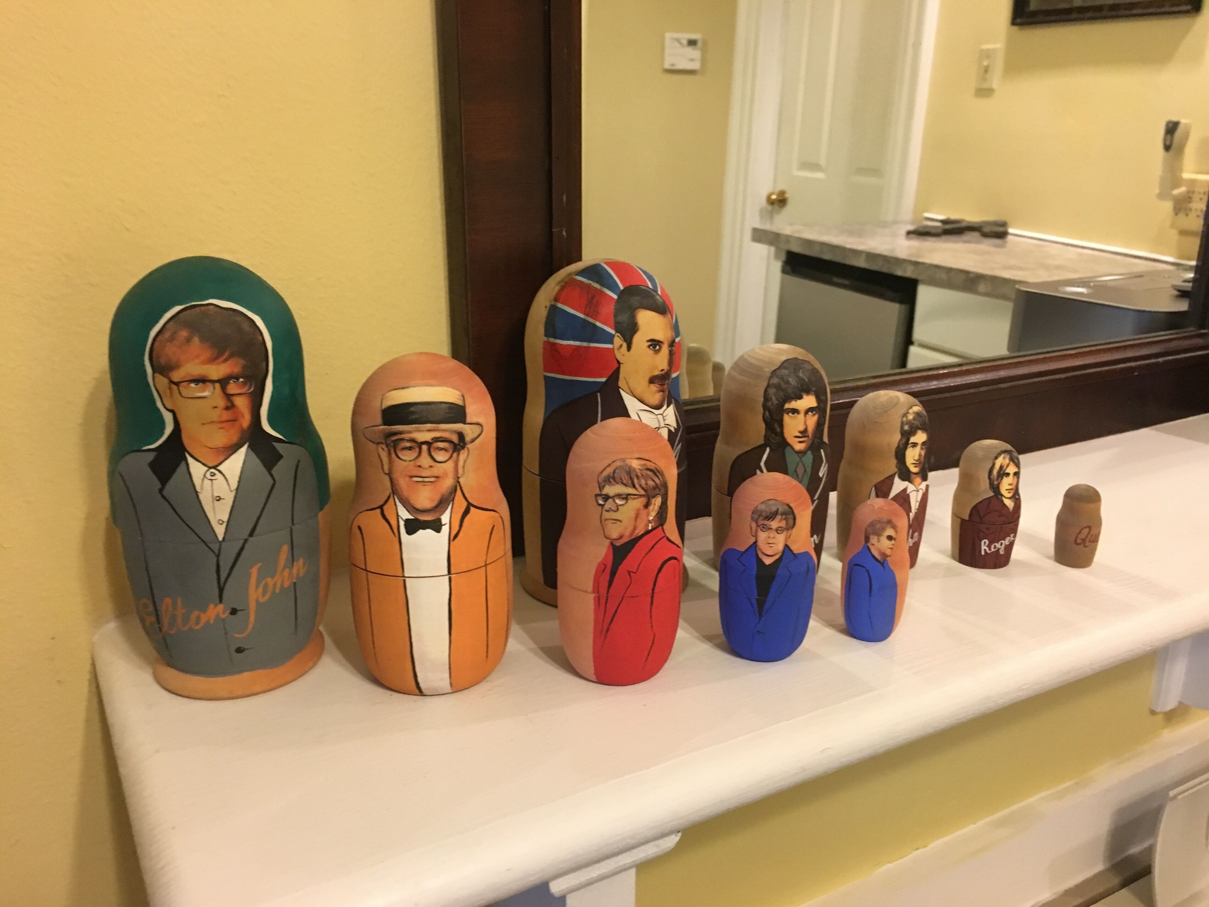 Eine Reihe bunte Holzfiguren auf einem weißen Regal vor einer gelben Wand.