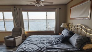 Een slaapkamer met een groot bed, een stoel, een plafondventilator, een raam met uitzicht op het water en een lamp.