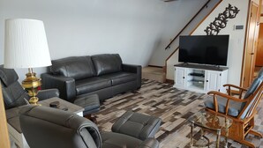 Een woonkamer met een bank, fauteuil en een glazen salontafel. Een televisie is bevestigd aan een kast, en er is een trap op de achtergrond te zien.