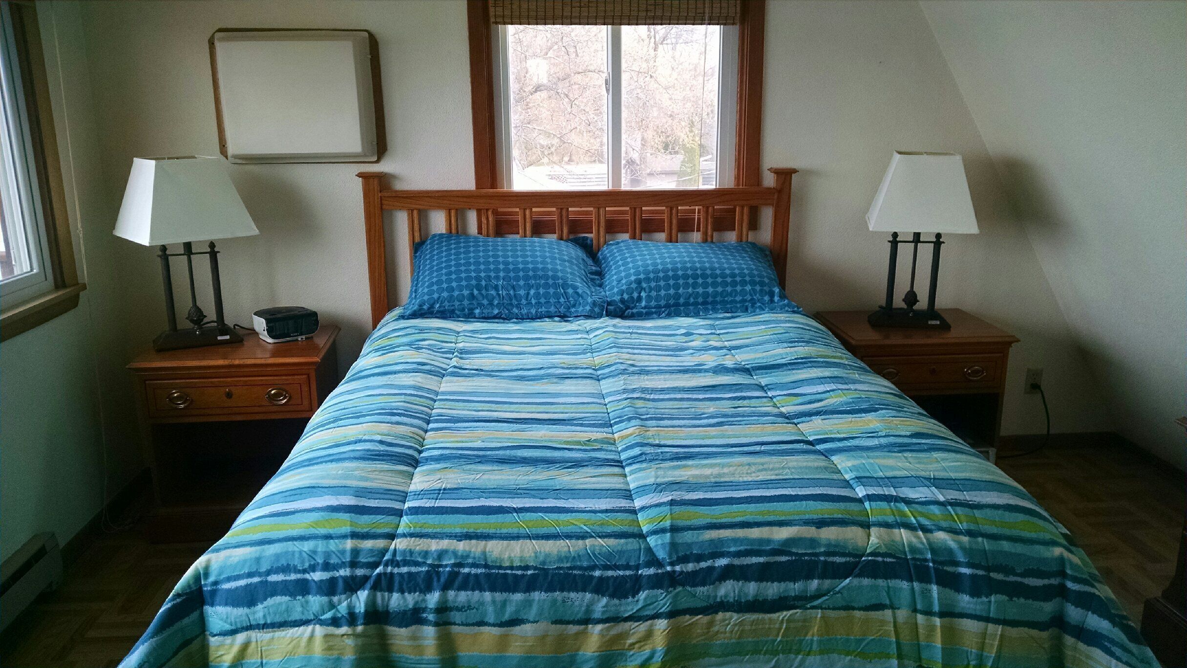 Un dormitorio con una cama, dos mesitas de noche, dos lámparas, una ventana y un cuadro colgado en la pared.