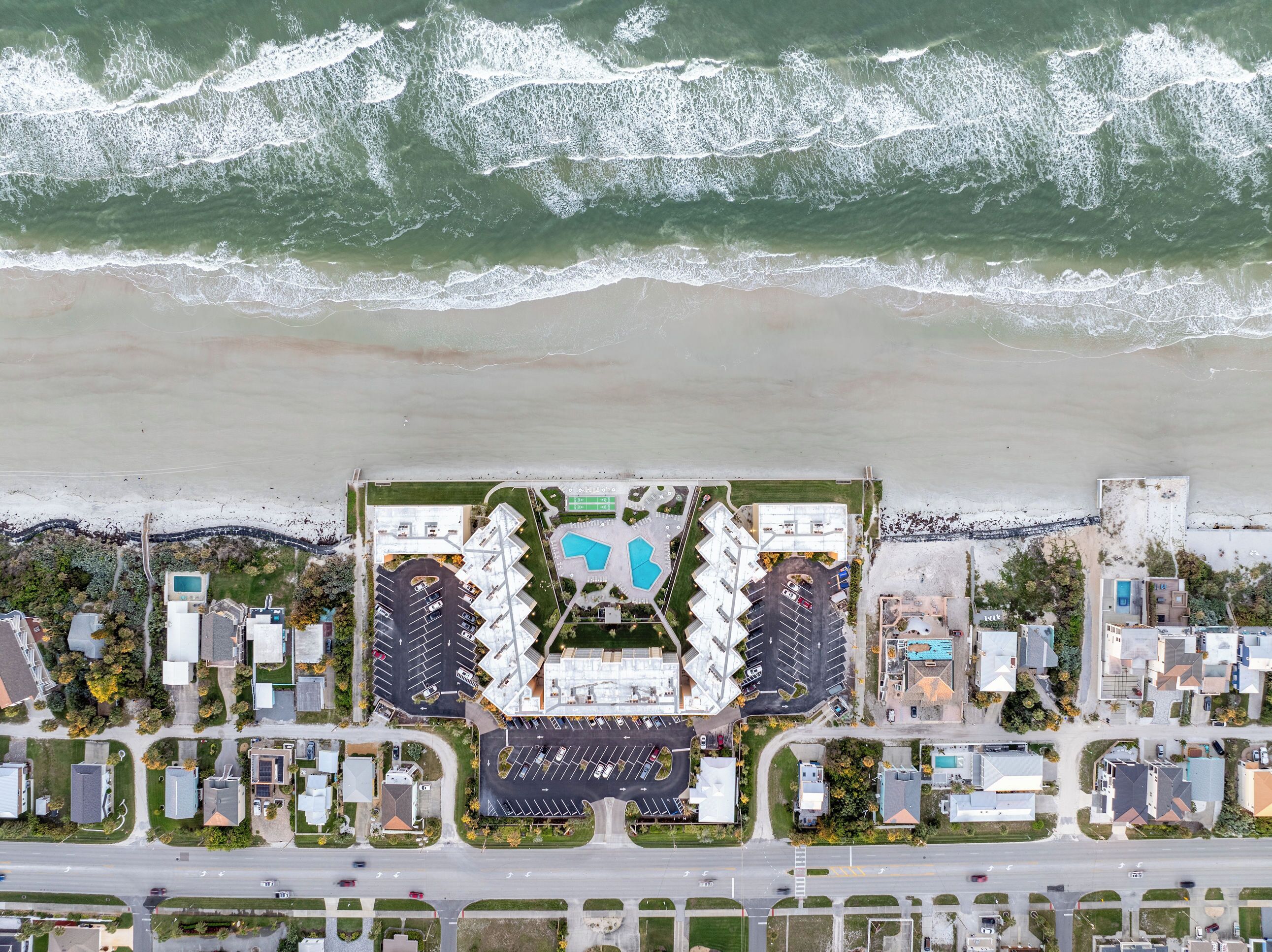 Vista aérea de un hotel costero con un estacionamiento central, varias edificaciones y una playa al fondo.