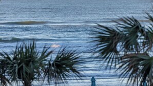 Vista del océano con olas, palmeras y una sombrilla azul.