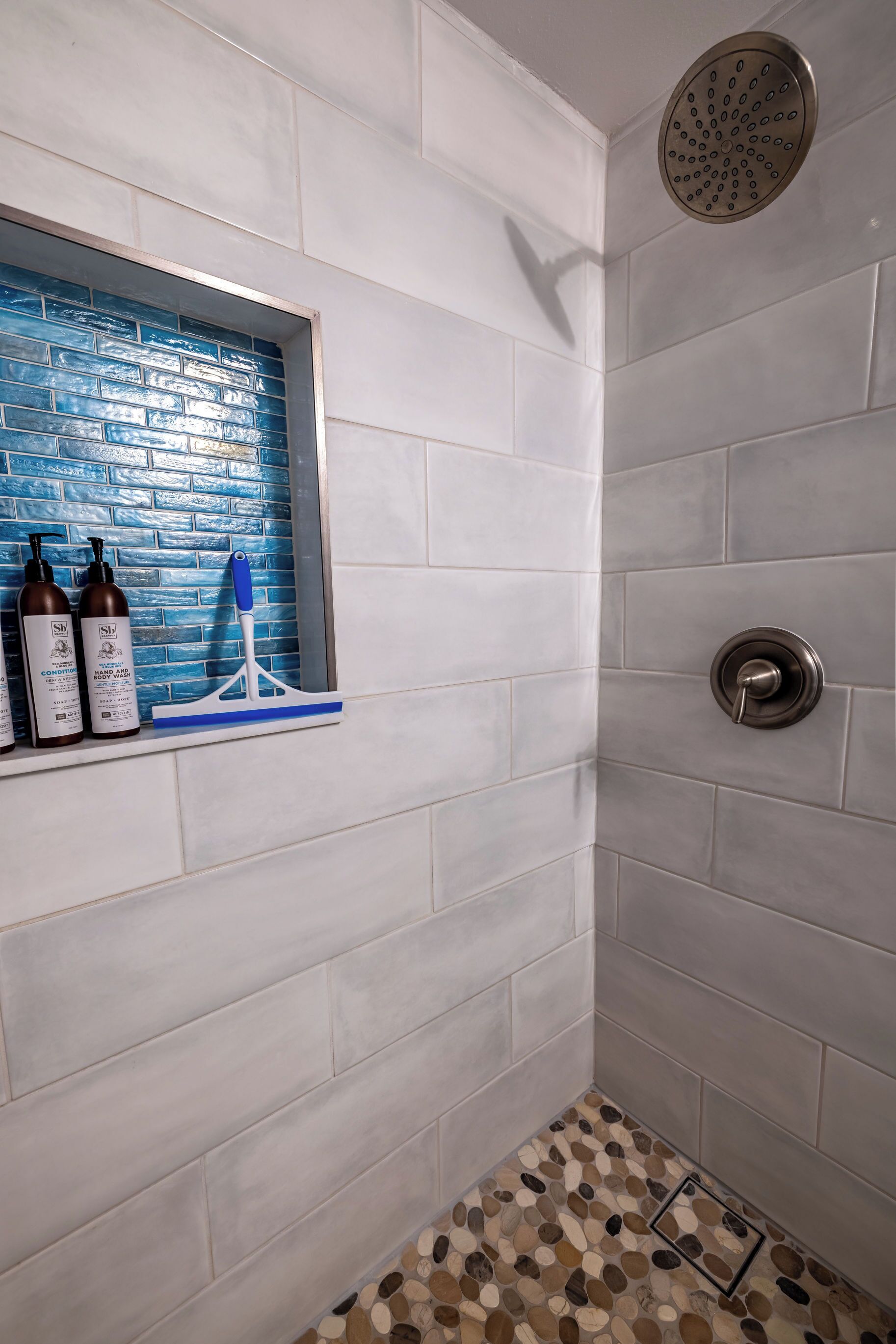 Un baño con azulejos blancos, un repisa de ventana con azulejos azules, un cabezal de ducha y un suelo con diseño de guijarros.