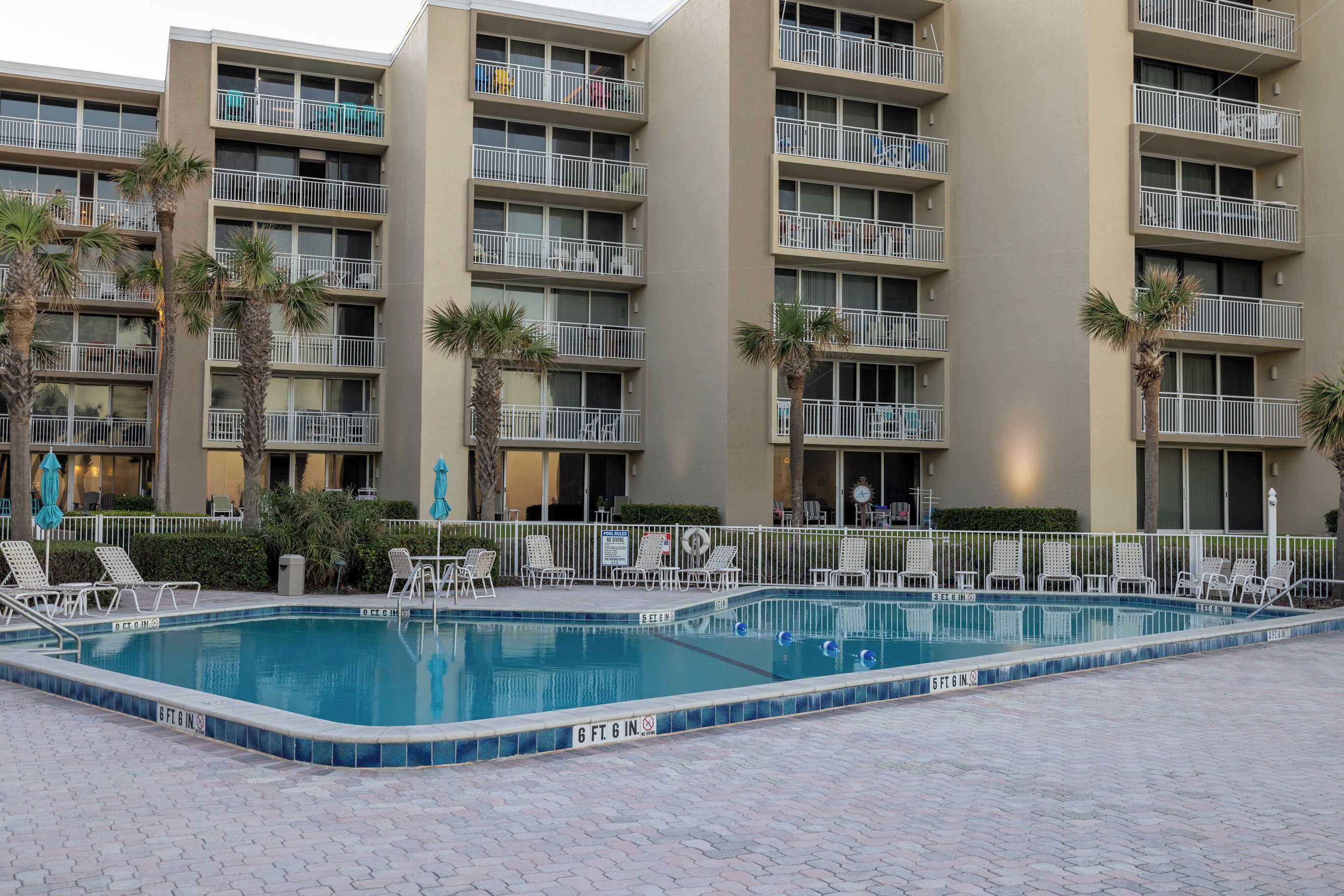 A swimming pool with lounge chairs and palm trees, surrounded by a multi-story building with balconies.