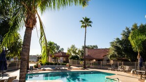 A swimming pool with lounge chairs and palm trees, surrounded by a fenced area and a building with a red-tiled roof.