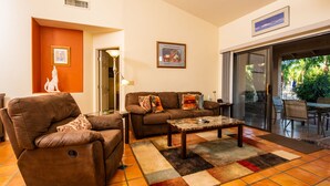 A living room with a brown sofa, a brown armchair, a coffee table, and a patterned rug. There is a door leading to a balcony with outdoor furniture.