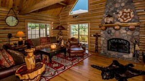 A log cabin interior with a stone fireplace, wooden furniture, and a large window overlooking greenery. - Luxury Log Home with Hot Tub on 7+ Acres Featured In Log Home Digest. Sleeps 10 (Pagosa Springs)