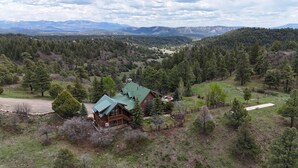 A log cabin with a green roof, surrounded by a forested area and mountains in the background. - Luxury Log Home with Hot Tub on 7+ Acres Featured In Log Home Digest. Sleeps 10 (Pagosa Springs)