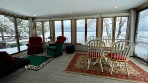 A room with large windows, a dining area with white chairs, a red armchair, and a green chair.