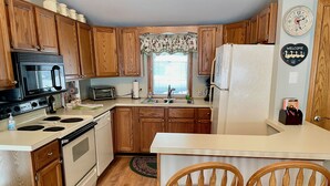 A compact kitchen with wooden cabinets, a microwave, stove, and a refrigerator. There are two wooden chairs with red cushions.