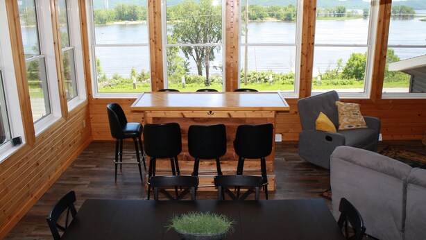 Interior de madera con una mesa de comedor, sillas y un sofá. Amplias ventanas ofrecen vistas a un lago y vegetación.