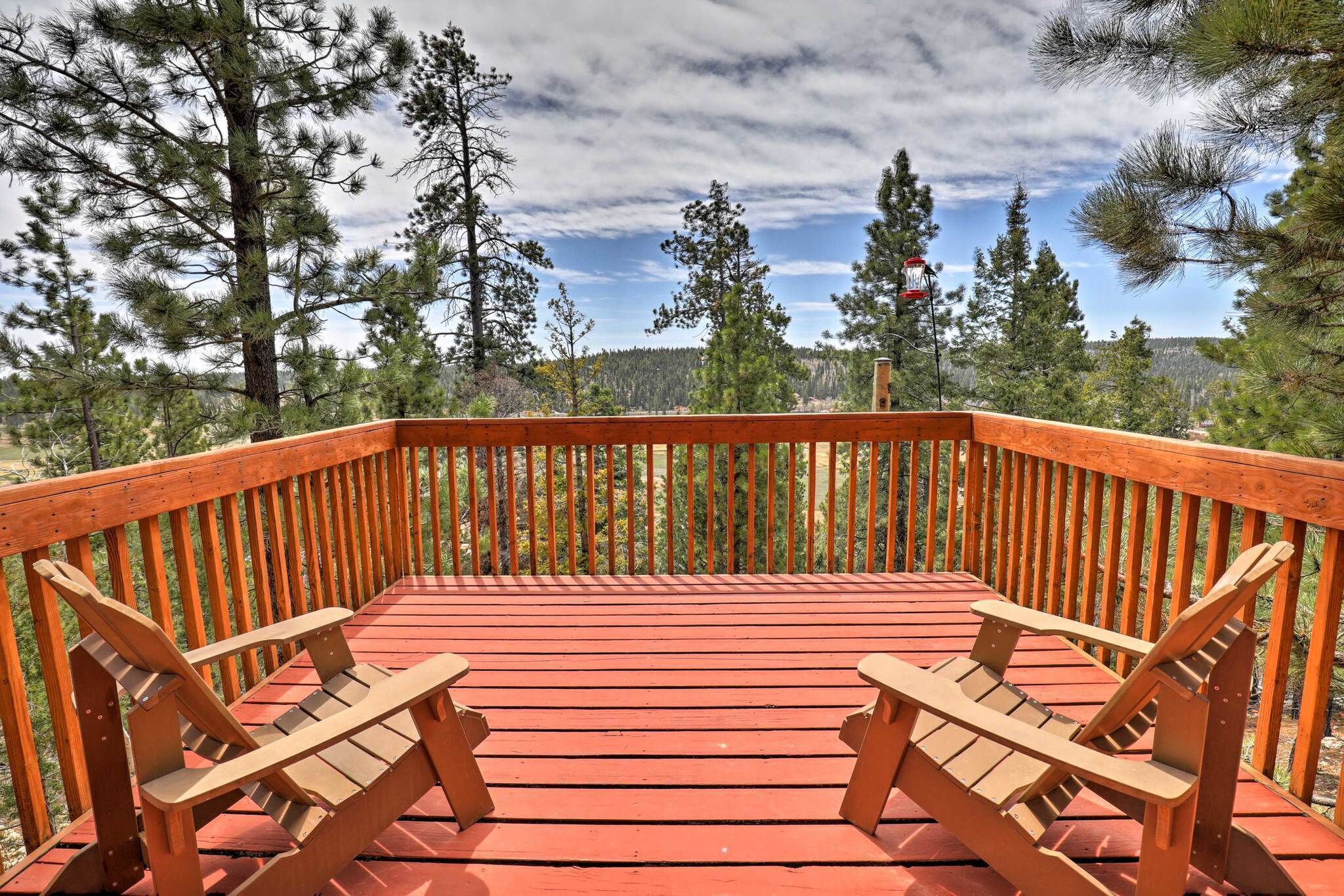 A wooden deck with two Adirondack chairs, surrounded by a railing, overlooking a forested area.