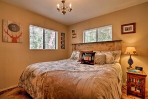 A bedroom with a large bed, two windows with blinds, a chandelier, a bedside table with a lamp, and wall art. - Fire Pit + Stunning Views ~ 2BD 1 BTH Mountain Cabin - Quiet, Dead End Lane (Duck Creek Village)