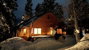 A wooden cabin with a porch, surrounded by snow-covered ground and trees.