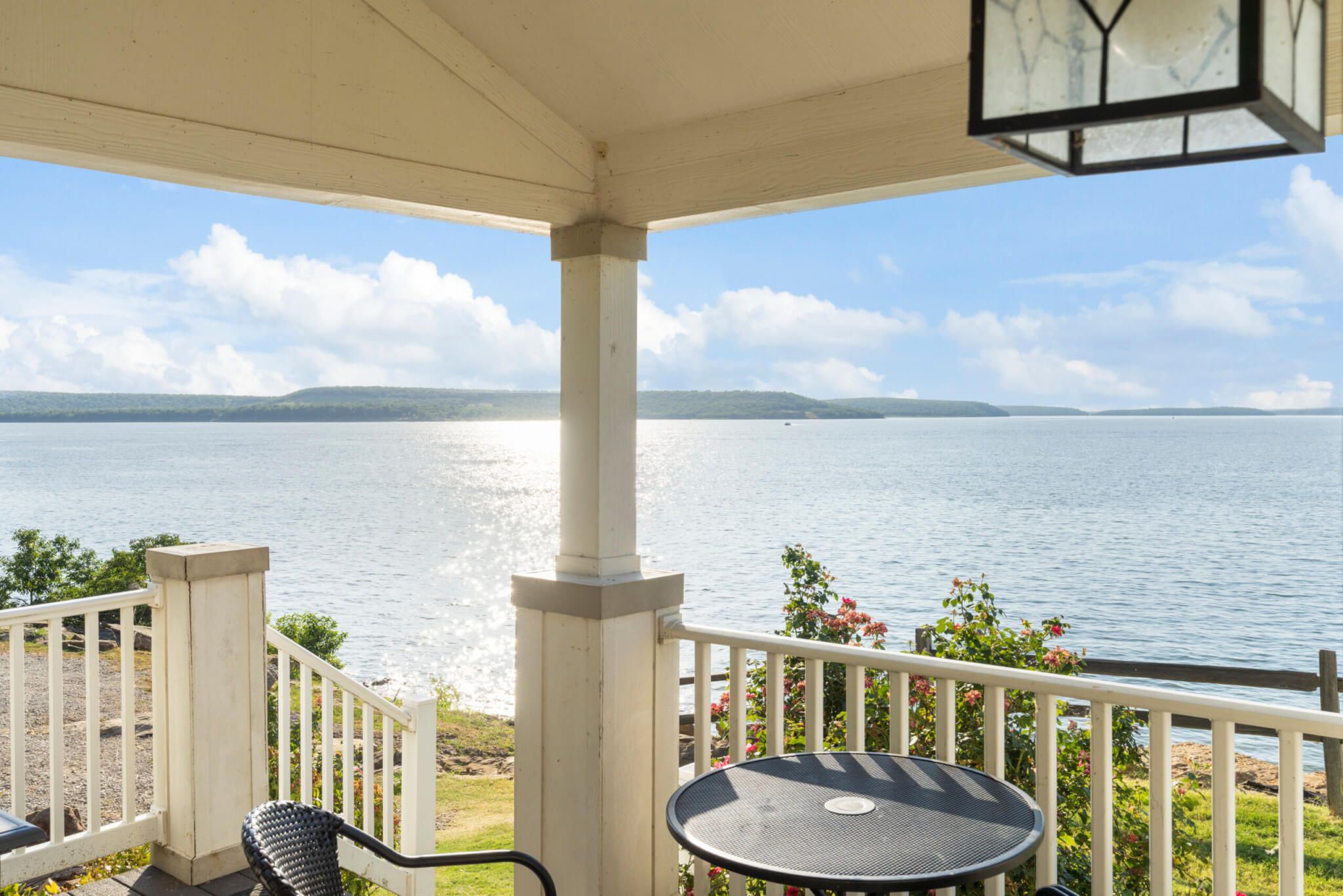 A covered outdoor area with a view of a large body of water, a white railing, and a round table.