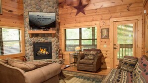 A cozy cabin interior with a stone fireplace, a television mounted above it, and a comfortable seating area with a sofa and armchairs. - Hillbilly Hotel, Great Resort, Great Game Room (Sevierville)