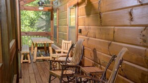 A wooden cabin with a porch featuring a table and chairs, large windows, and a view of greenery. - Hillbilly Hotel, Great Resort, Great Game Room (Sevierville)