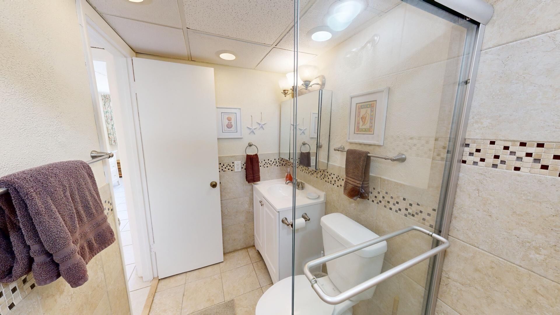 Une salle de bain avec une douche à parois de verre, des toilettes blanches, un lavabo avec un miroir et un porte-serviettes avec une serviette marron.