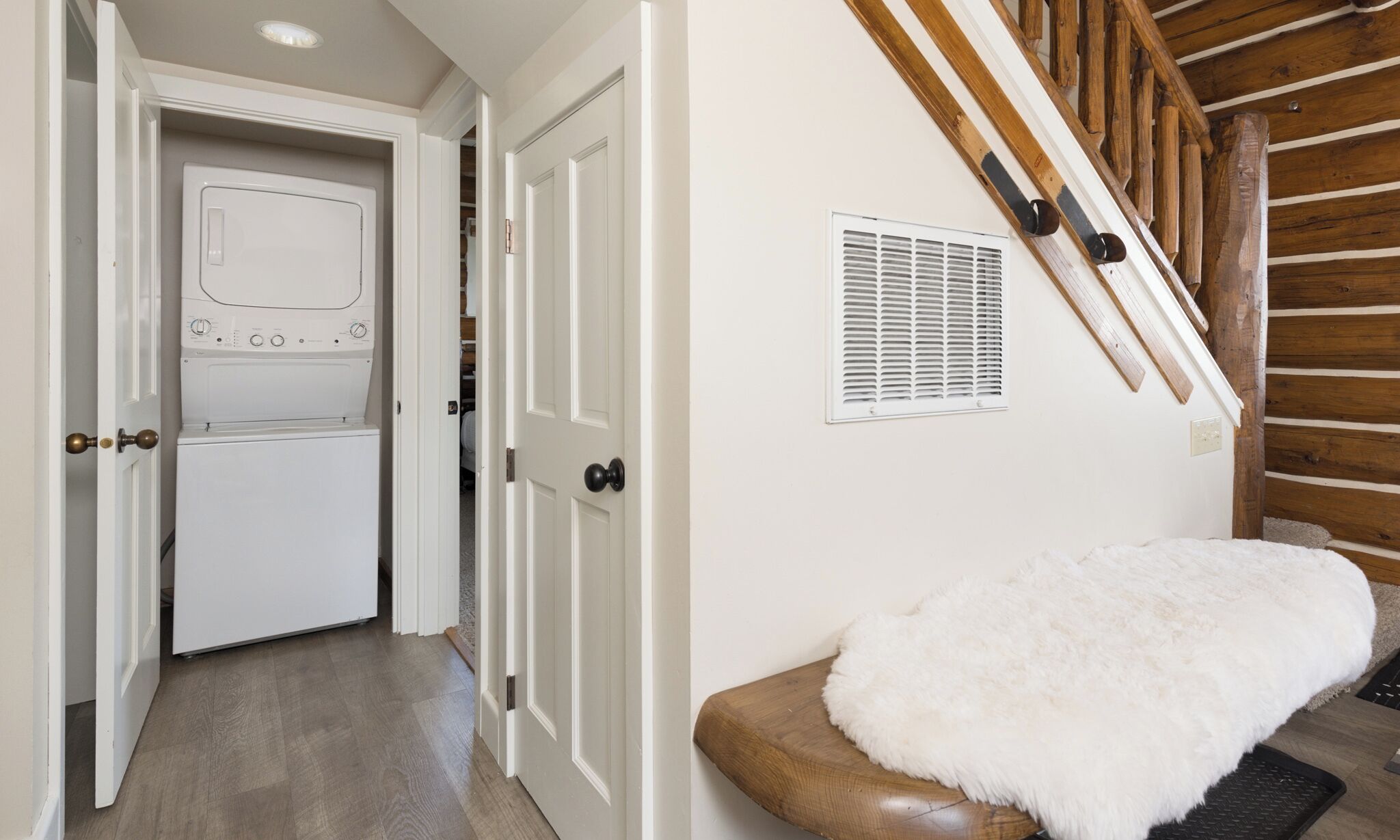 A hallway with a laundry room, wooden staircase, and a white fluffy rug on a wooden bench.