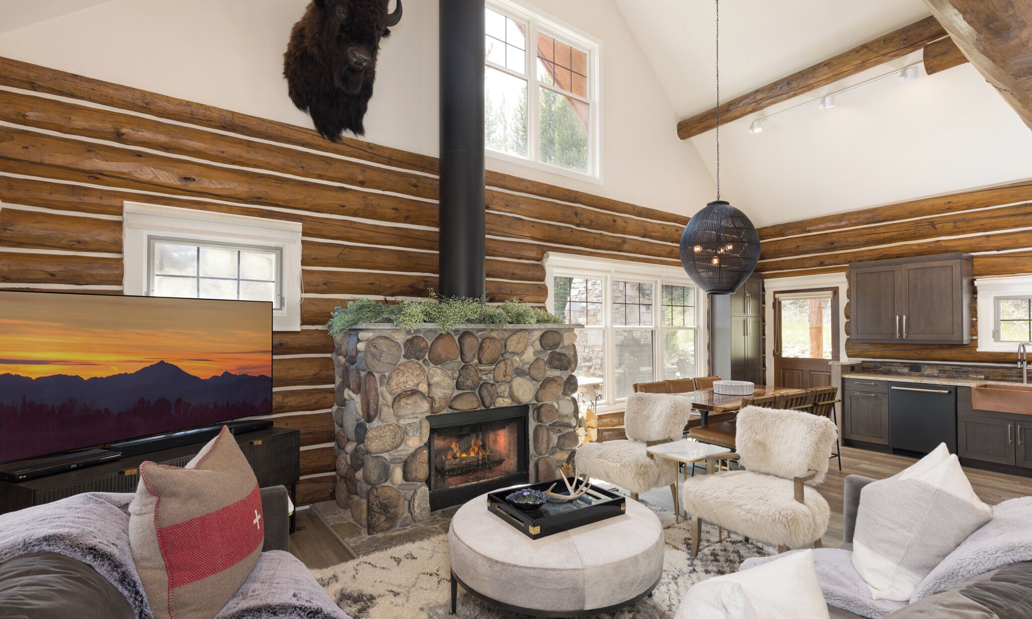 A log cabin interior with a stone fireplace, wooden walls, a dining area, and a kitchen.