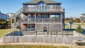 A multi-story wooden building with balconies, a central staircase, and a fenced area in front. - Oceanview Dog Friendly Home w/ Private Pool, Hot Tub, and Beach Access Nearby (Rodanthe)