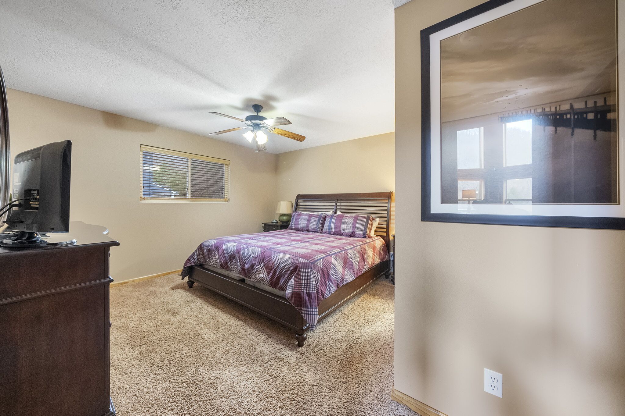 A bedroom with a bed, a ceiling fan, a dresser, a window with blinds, and a framed picture on the wall.