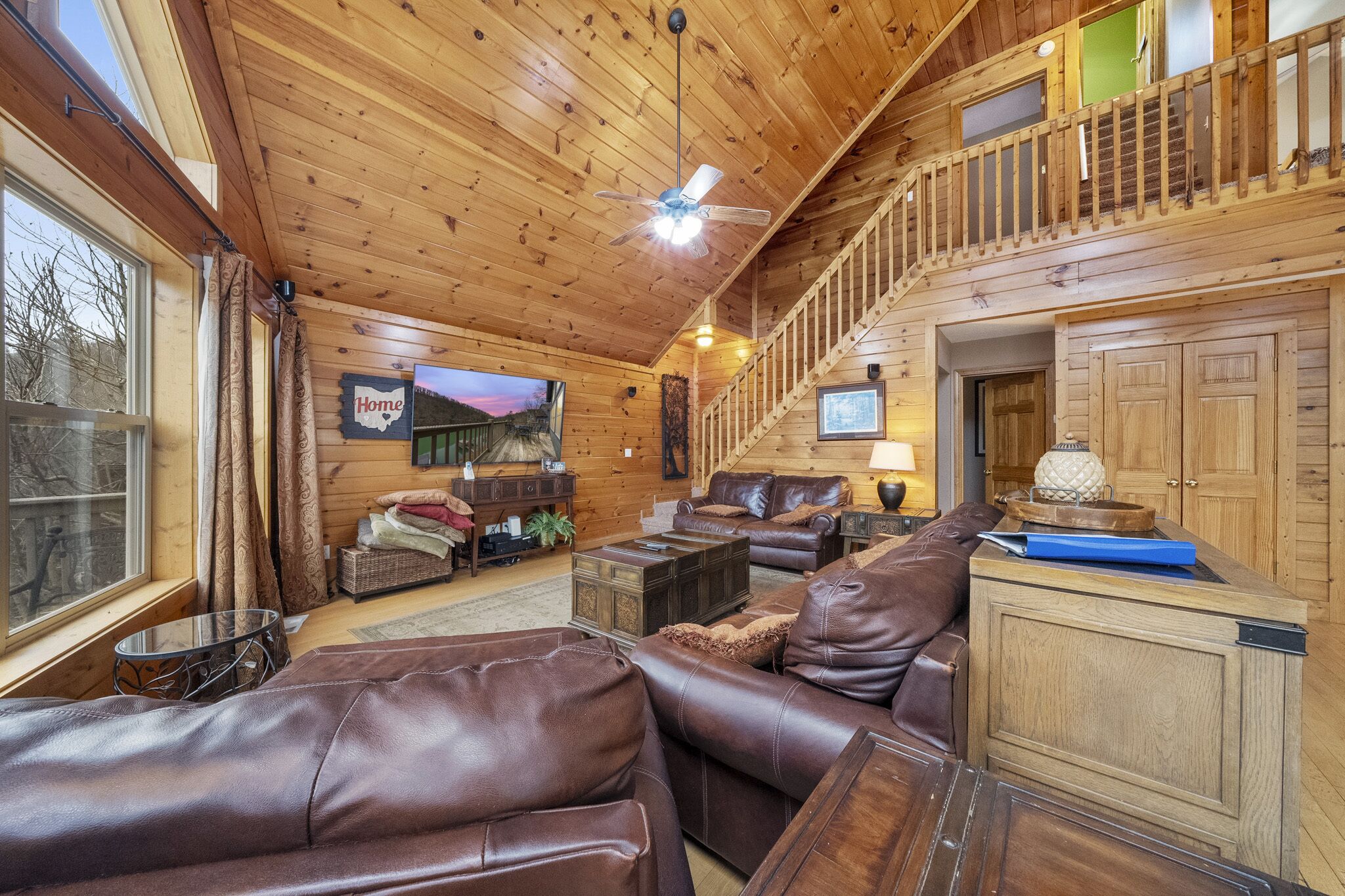 A cozy cabin interior with a wooden ceiling, leather sofas, a television, and a staircase to an upper level.