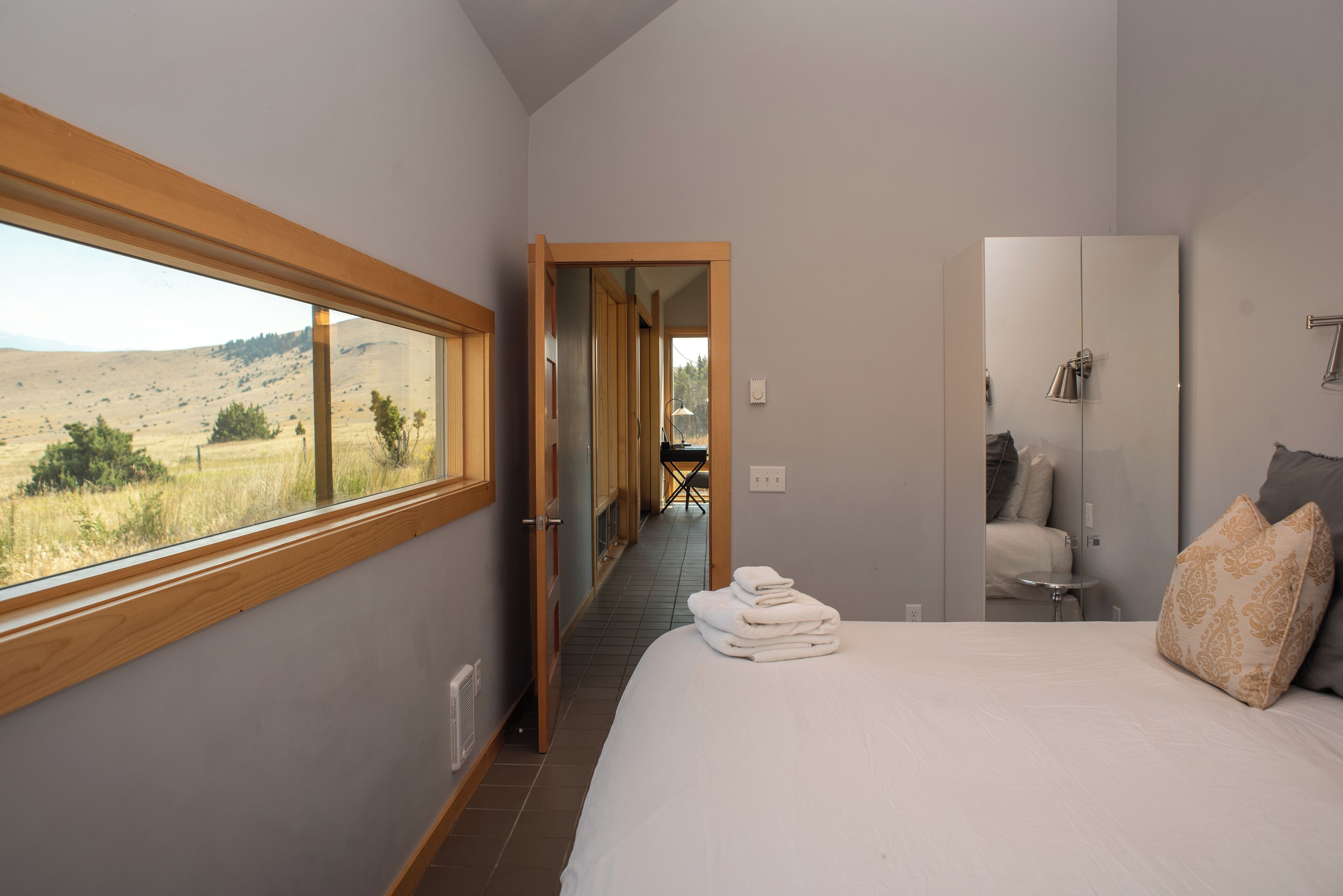 Un dormitorio con un ventanal grande, una cama con toallas ordenadas, un armario y vistas a un paisaje rural.