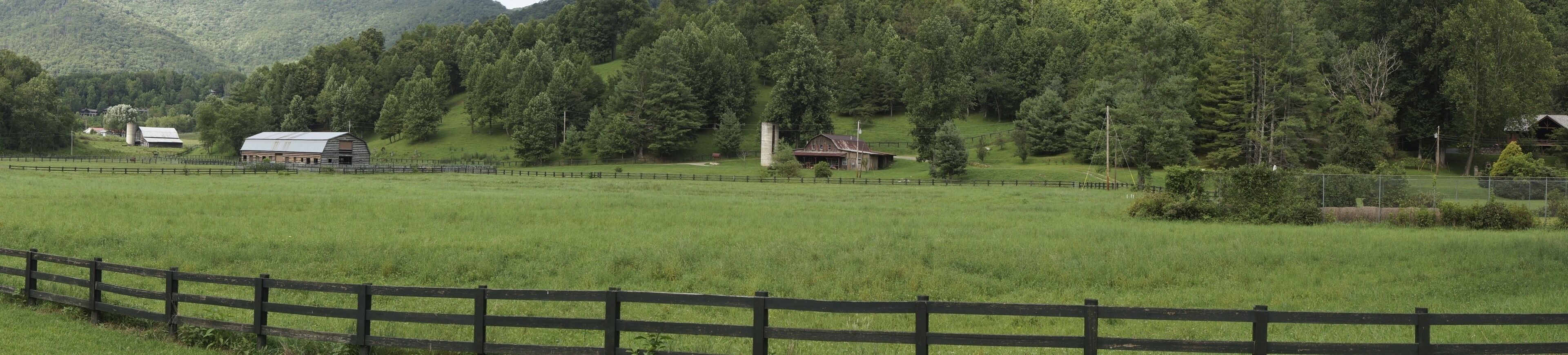Paisagem rural com cerca de madeira, um celeiro e uma casa rodeada por árvores e colinas.