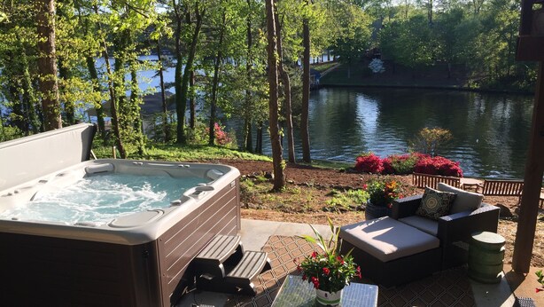 A hot tub with a view of a lake, surrounded by outdoor furniture and plants. - Amazing Views at Dragonfly! HOT TUB! Kayaks, BOUNCER w Slide, SUP's, Fire Pit!  (Bracey)