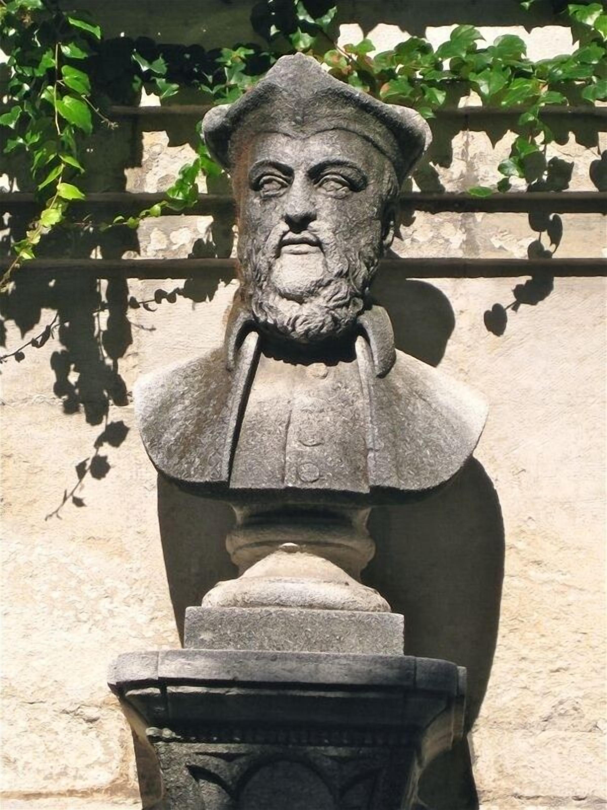A stone bust sculpture mounted on a pedestal, set against a stone wall with ivy.