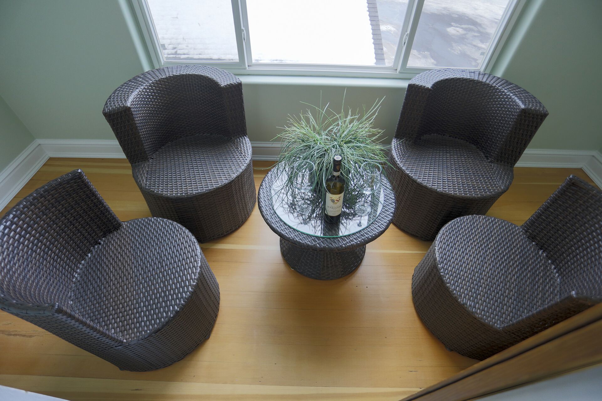 Une pièce comprenant quatre chaises en osier disposées autour d’une table dont le plateau est en verre, avec une bouteille et des verres.