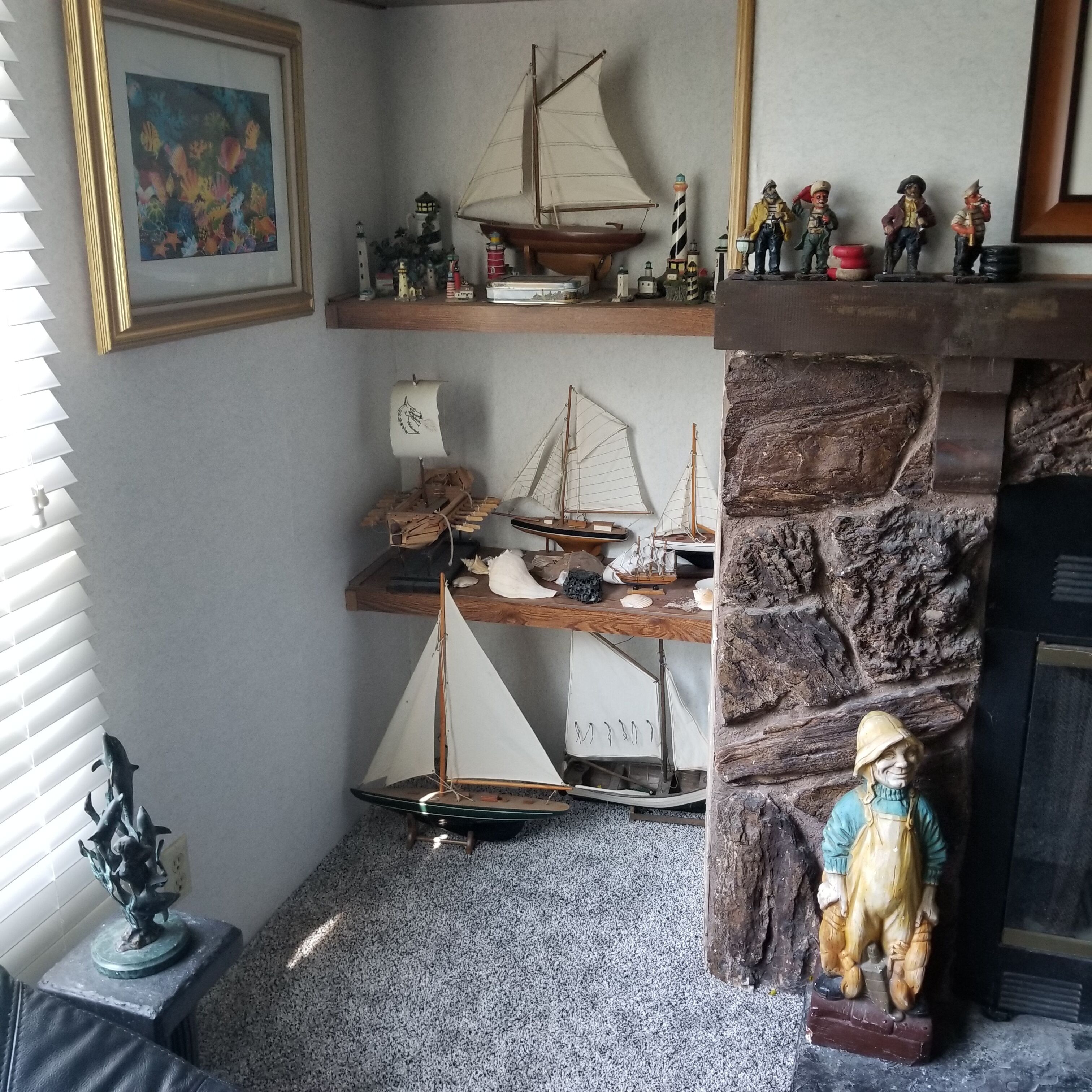 Een hoekzaal met een stenen openhaard, een houten plank met scheepsmodellen, een klein beeldje en een lijst met een schilderij.