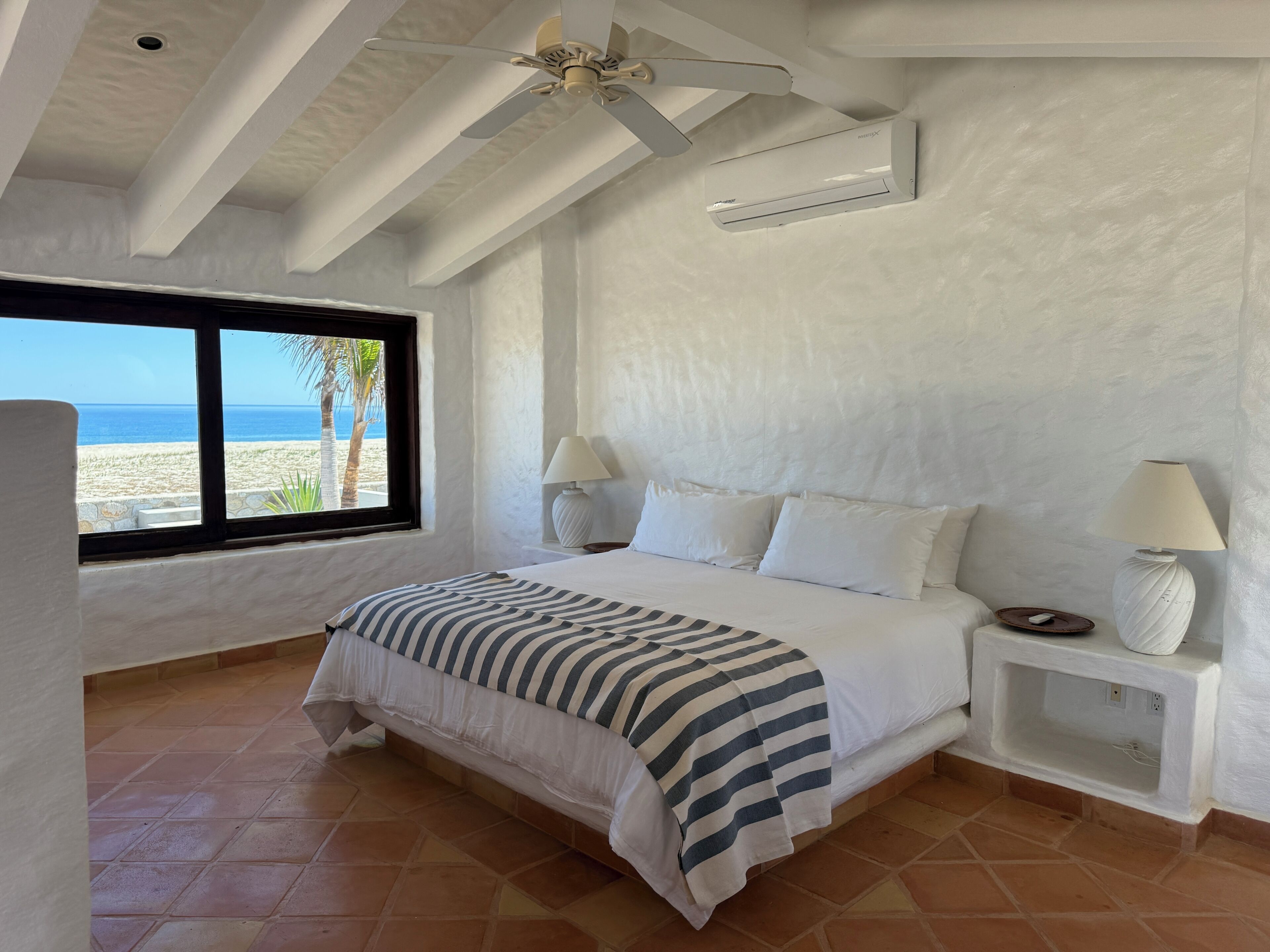 A bedroom with a bed, two bedside tables with lamps, a ceiling fan, an air conditioner, a window with a beach view, and a tiled floor.