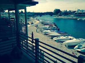 A beach with rows of boats docked along the shore, a bridge in the background, and a building with a balcony overlooking the beach.