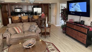 Una sala de estar con un sofá de cuero marrón, una mesa de centro con un control remoto, un televisor y una cocina al fondo.
