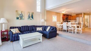 Ein Wohnzimmer mit einem Ecksofa, einem weißen Couchtisch und einem Essbereich mit Tisch und Stühlen. An den Wänden befinden sich Gemälde und es gibt einen Deckenventilator.